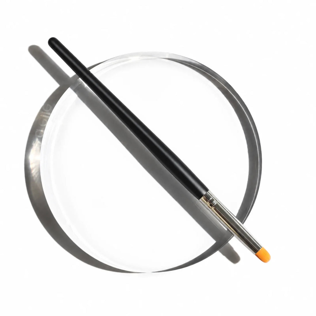 Black-handled Lomansa lash lift applicator brush with a silver ferrule and orange tip resting on a clear round tray.