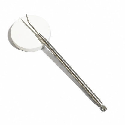  Lomansa silver dual-ended lash lift separator beside a white circular prop, showing the angled hook and fine comb.
