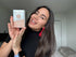 Smiling woman with long dark hair holding a peach and white LOMANSA lash and brow tint kit box beside her face.